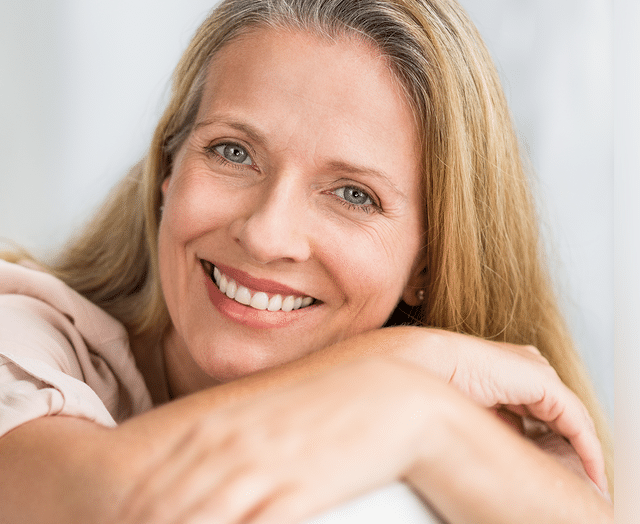 Smiling woman with healthy teeth resting chin on hand against white background.
