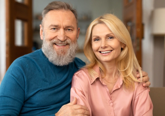 Smiling middle-aged couple posing together in home interior setting for dentures website.