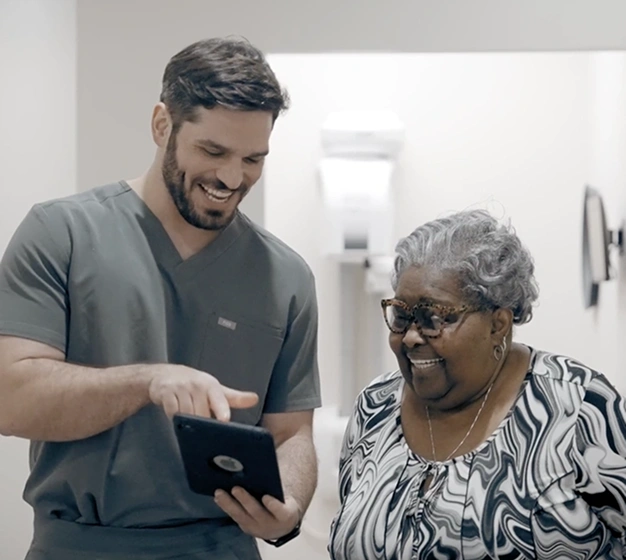 Young male dental professional showing tablet to smiling elderly woman in clinical setting.
