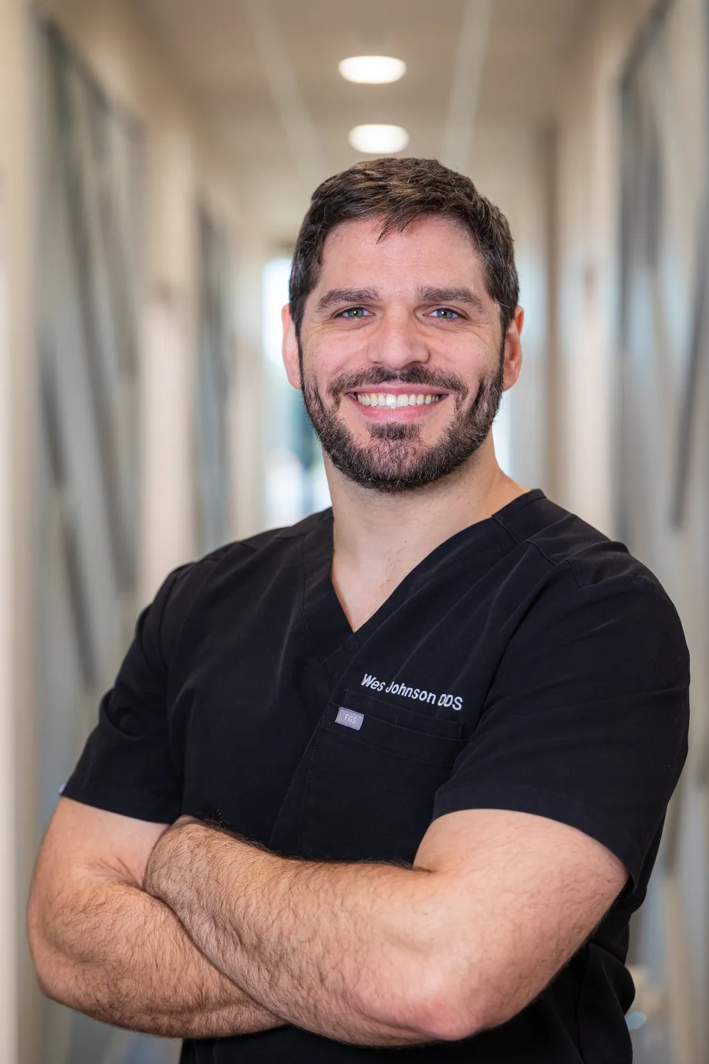 Professional male dentist smiling in dental office hallway wearing black scrubs