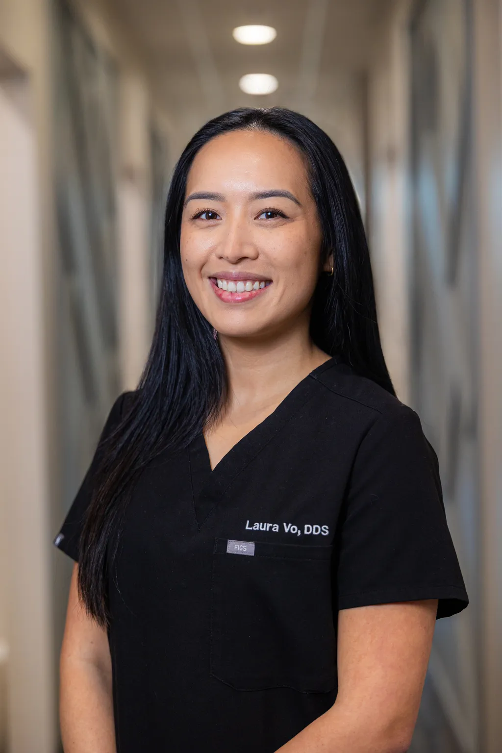 Professional woman in black scrubs smiling in modern dental office hallway.