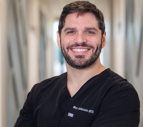 Professional headshot of smiling male dentist wearing black scrubs in clinical office setting.