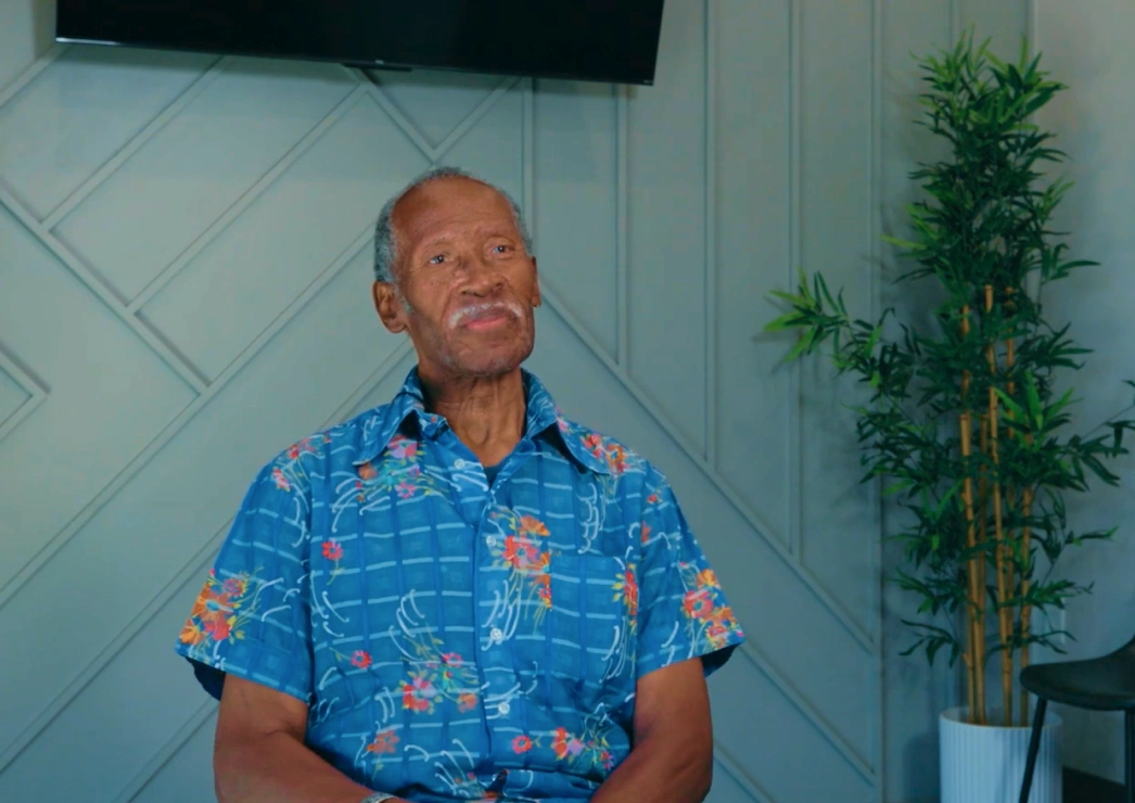 Man in blue patterned shirt standing indoors with modern white wall and potted plant behind him.