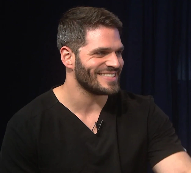 Man smiling during interview or presentation against dark background, wearing black v-neck shirt.